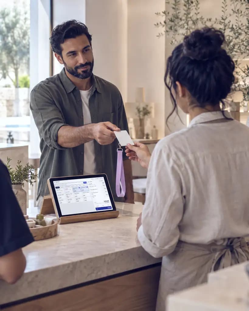 An owner hands a staff card to a new cashier across the counter; the till tablet sits between them.